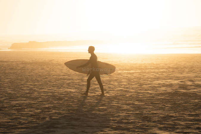 solo traveller carrying surfboard and walking through sunset beach