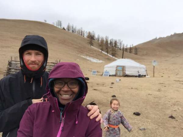 travelling couple in Mongolia smiling at the camera while wearing raincoats, local child running through desert and ger camp in background