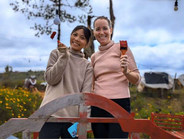 travel-solo-or-with-friend-comparison two workawayers smiling at camera while holding paintbrushes small woodwork project volunteering outdoor task experience